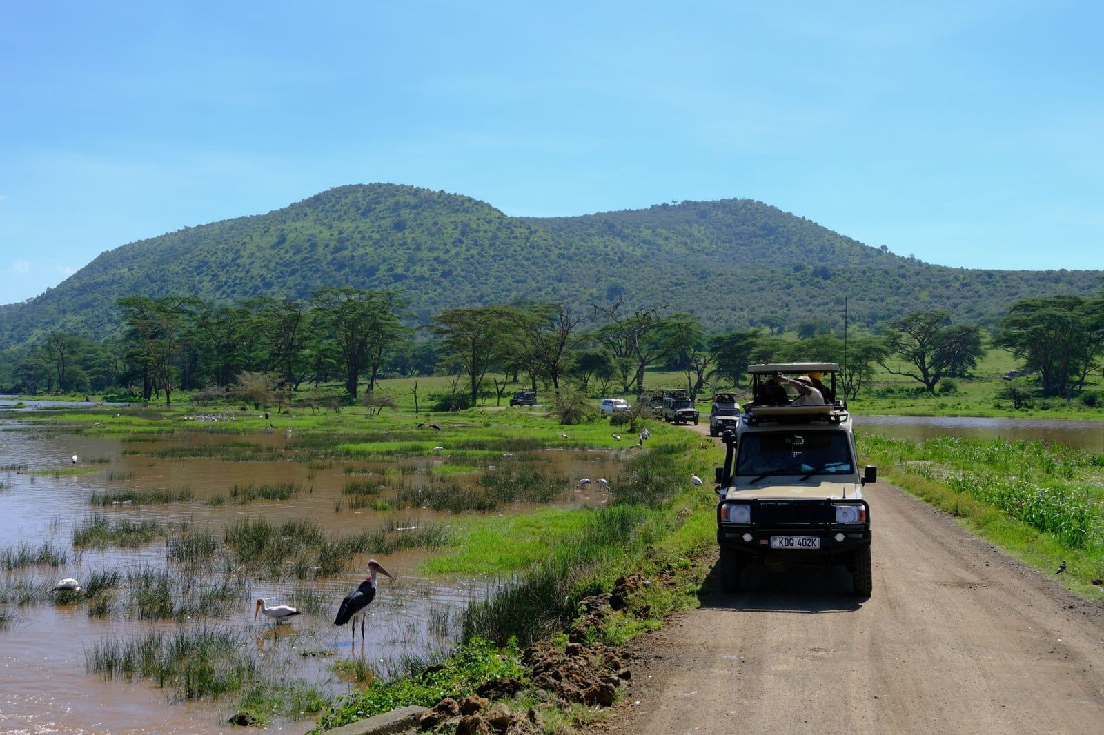 A safari vehicle drives along a dirt road near the marshy shores and green hills of Lake Nakuru.